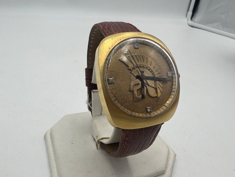 Puede incluir: Un reloj de pulsera dorado con una correa de cuero marr&oacute;n. La esfera del reloj presenta una esfera romana dorada con agujas doradas de hora y minuto. La esfera del reloj tambi&eacute;n presenta una imagen dorada de un casco de guerrero.