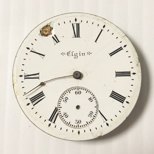 May include: White pocket watch face with black Roman numeral hour markers and black minute markers. The watch face has a small, white sub-dial with black markings for seconds. The watch face is labeled "Elgin".
