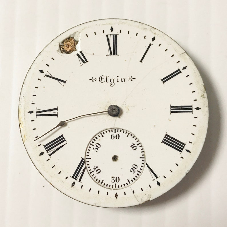 May include: White pocket watch face with black Roman numeral hour markers and black minute markers. The watch face has a small, white sub-dial with black markings for seconds. The watch face is labeled "Elgin".