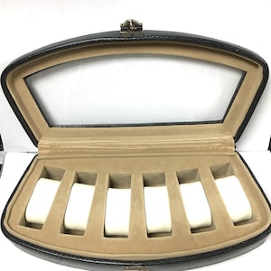 May include: Black leatherette watch display case with a clear glass top. The interior features six white watch cushions set against a tan felt lining. A silver-tone clasp secures the case.