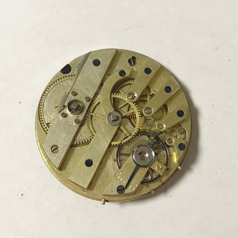 May include: A detailed view of a vintage watch movement, revealing the complex arrangement of gears, levers, and springs. The mechanical parts are set on a circular, gold-coloured base, with silver-coloured metal supports. The image highlights the precision and complexity of the watch's internal mechanisms.