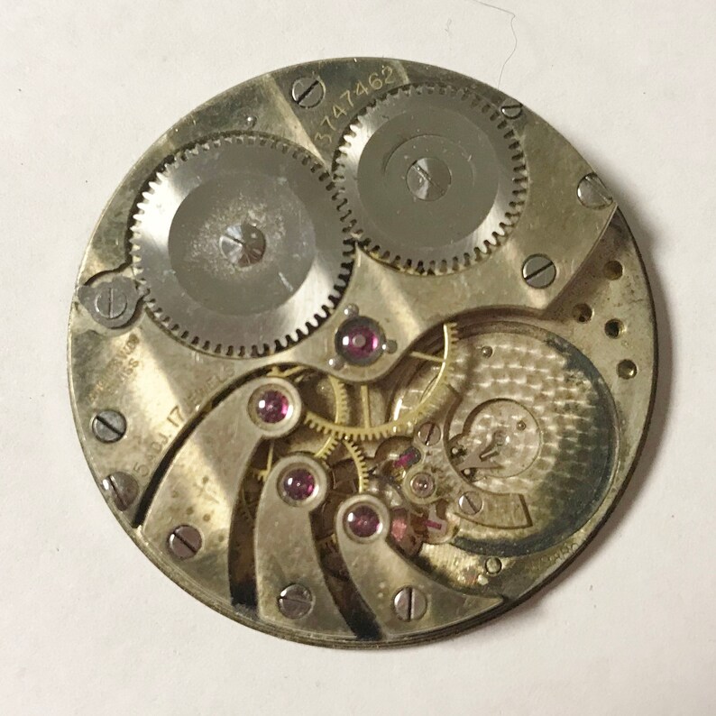 May include: Close-up of a silver watch movement with a gold-colored gear train and several ruby jewels. The movement is marked with the numbers "3747462" and "5817".