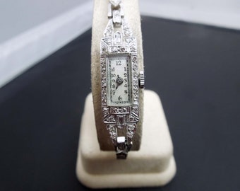 Op de afbeelding: Een zilveren polshorloge met een rechthoekige wijzerplaat en diamant accenten. Het horloge heeft een witte wijzerplaat met zwarte Romeinse cijfers als uurmarkeringen en zwarte wijzers.