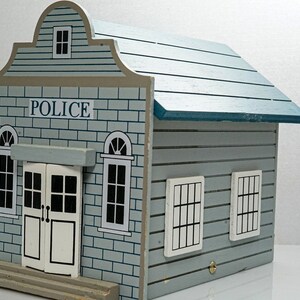 May include: A blue wooden toy police station with a sign that reads "POLICE" and windows with black frames.