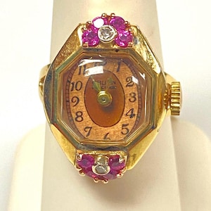 May include: A gold-toned watch ring with a brown watch face and gold hands. The ring features a decorative setting with pink gemstones and a single diamond. The watch ring is on a white background.