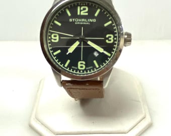 May include: A Stuhrling Original wristwatch with a round silver-tone metal case, a black dial featuring luminous numerals at 12, 3, 6, and 9, luminous hands, a date window at the 4 o'clock position, and a brown leather strap with visible stitching.