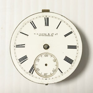 Op de afbeelding: Witte zakhorlogewijzerplaat met zwarte Romeinse cijfers voor de uren en zwarte minutenmarkeringen. De wijzerplaat van het horloge heeft de tekst "W. E. WATTS & CO. LD. STONEHOUSE" erop gedrukt.