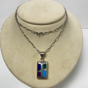 Puede incluir: Un collar de cadena de plata con un colgante rectangular con piedras de turquesa, malaquita y púrpura incrustadas.