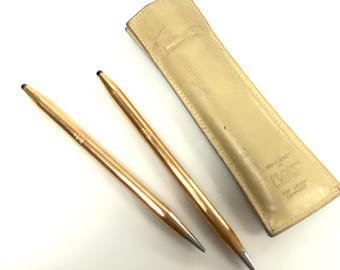 May include: Two gold-toned Cross pens with a leather case. The case is beige and has the words "PEN POCKET" and "CROSS" printed on it.