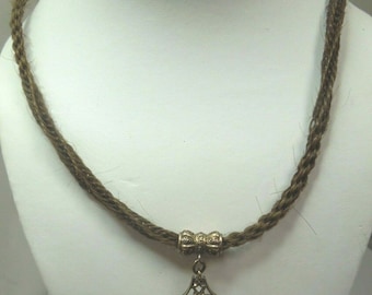 Puede incluir: Un collar de cordón trenzado marrón con un colgante dorado y una cuenta decorativa. El colgante tiene forma de sello con detalles intrincados.