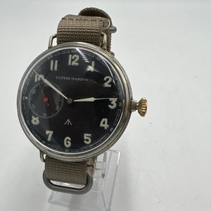 May include: A black-faced wristwatch with white numerals and hands. The watch has a silver case and a beige canvas strap. The watch face reads "ULYSSE NARDIN".
