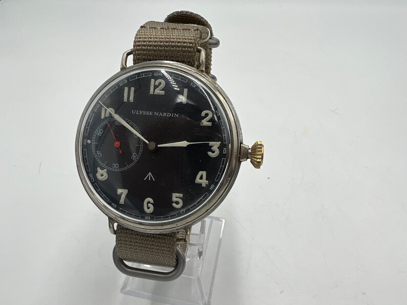 May include: A black-faced wristwatch with white numerals and hands. The watch has a silver case and a beige canvas strap. The watch face reads "ULYSSE NARDIN".