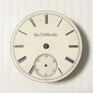 May include: White pocket watch face with black Roman numeral hour markers and black minute markers. The watch face has the text "Elgin National Watch Co." printed on it.