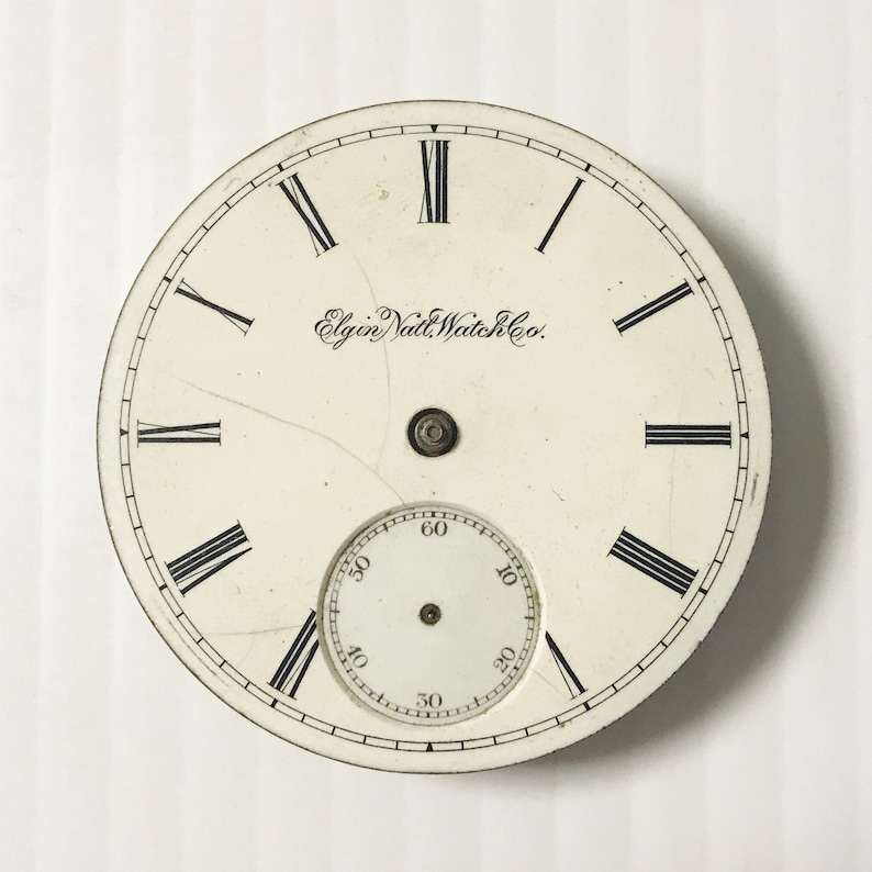 May include: White pocket watch face with black Roman numeral hour markers and black minute markers. The watch face has the text "Elgin National Watch Co." printed on it.