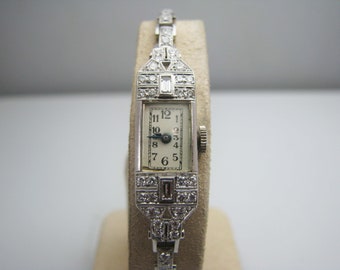 Könnte beinhalten: Eine vintage Armbanduhr mit Diamantenbesatz, weißem Zifferblatt und schwarzen römischen Ziffern. Die Uhr hat ein silberfarbenes Metallband.
