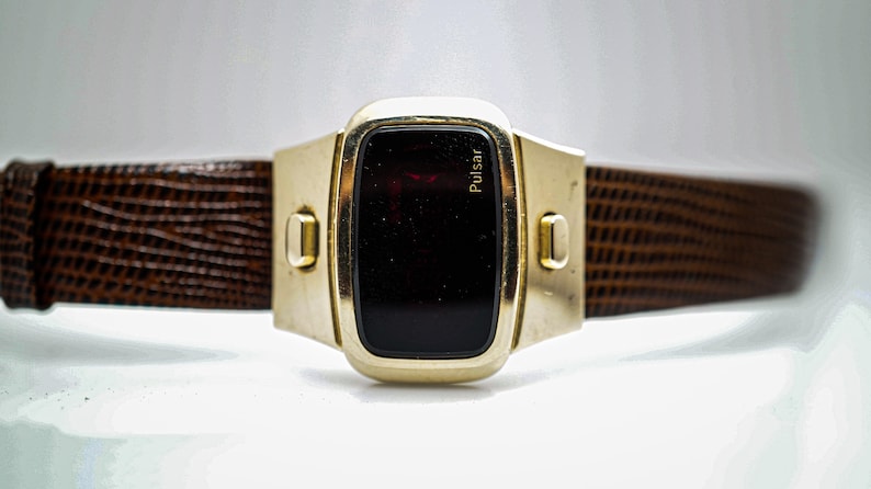 May include: A gold-toned Pulsar digital watch with a brown leather strap. The watch face is a dark red LED display.