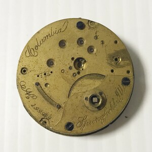 May include: Close-up of a vintage watch movement with a brass finish. The words "Columbia" and "Springfield Ill." are inscribed in cursive. The serial number "No. 199949" is also visible.