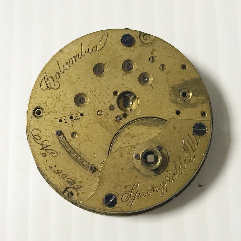 May include: Close-up of a vintage watch movement with a brass finish. The words "Columbia" and "Springfield Ill." are inscribed in cursive. The serial number "No. 199949" is also visible.