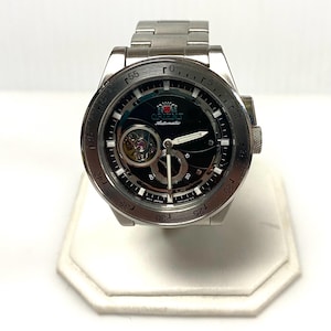 May include: A silver-toned Orient automatic wristwatch with a black face. The watch features a visible inner mechanism and a silver band. The face includes the Orient logo and the word "Automatic". The watch is displayed on a white stand.
