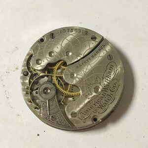 Op de afbeelding: Close-up van een zilveren zakhorloge-uurwerk met de inscriptie "American Waltham Watch Co." en "Safety Barrel" zichtbaar. Het uurwerk heeft het serienummer "10756912" bovenaan.