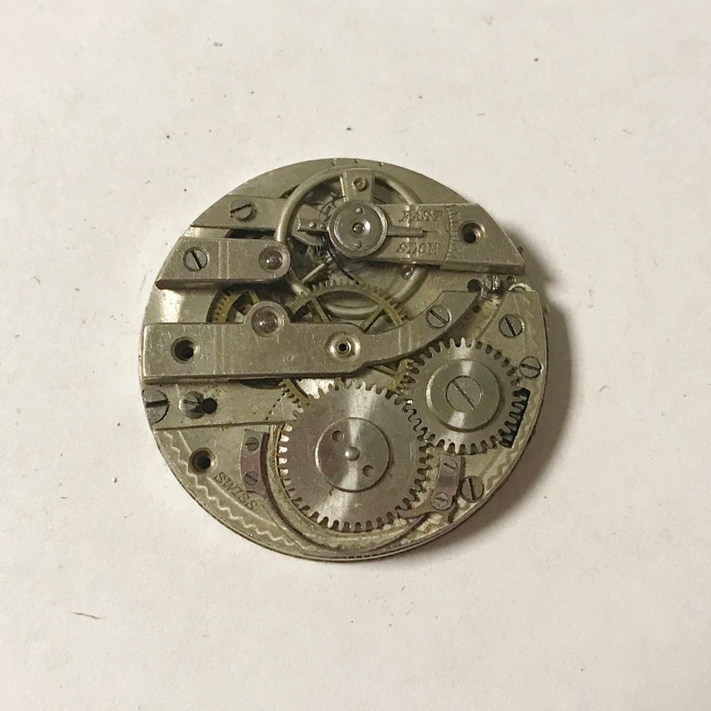 Puede incluir: Primer plano de un movimiento de reloj plateado con engranajes intrincados y una palanca de ajuste "R&Aacute;PIDO LENTO". El movimiento est&aacute; etiquetado como "SWISS".