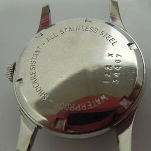 May include: The back of a silver wristwatch with the text "WATERPROOF - SHOCKRESISTANT - ALL STAINLESS STEEL" and the numbers "34407 122 X" engraved on the case.