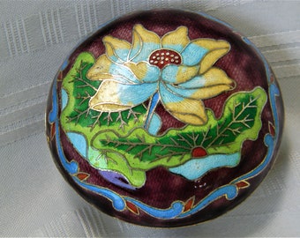 May include: A round, purple, cloisonné trinket box with a blue and yellow lotus flower design. The box is decorated with green leaves and a gold trim.