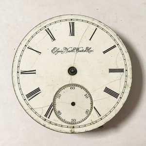 May include: A white watch face with black Roman numeral hour markers and black Arabic numeral minute markers. The watch face has a small, circular subdial with black Arabic numeral markers. The text "Elgin Natl. Watch Co." is printed on the watch face.