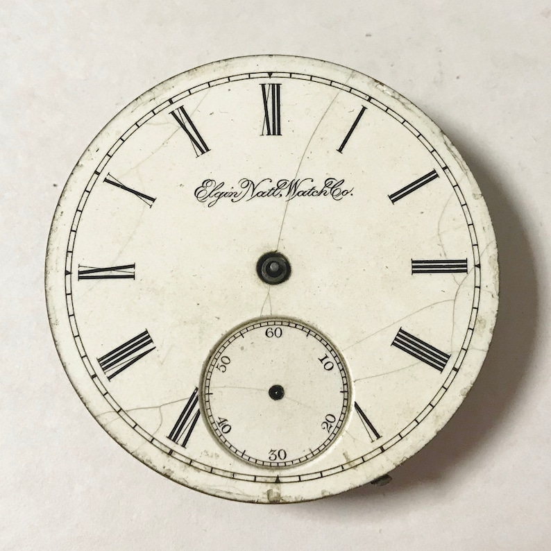 May include: A white watch face with black Roman numeral hour markers and black Arabic numeral minute markers. The watch face has a small, circular subdial with black Arabic numeral markers. The text "Elgin Natl. Watch Co." is printed on the watch face.