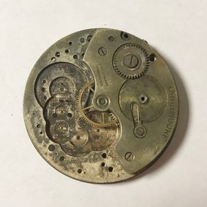 May include: Close-up of a silver watch movement with the text "SWISS  MADE  UNADJUSTED" and "LANGENDORFF" visible. The movement has several gears and a winding stem.