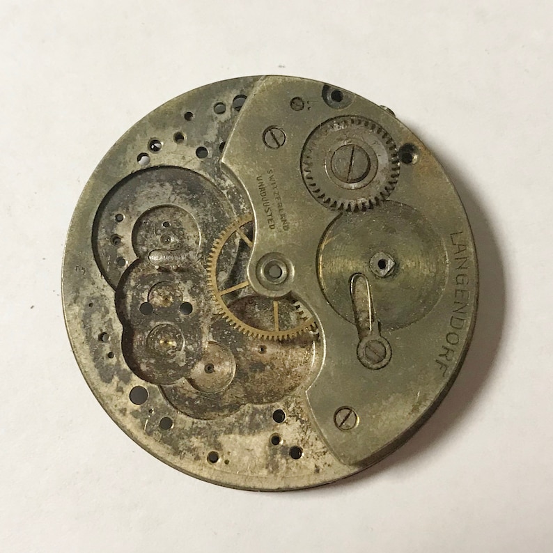 May include: Close-up of a silver watch movement with the text "SWISS  MADE  UNADJUSTED" and "LANGENDORFF" visible. The movement has several gears and a winding stem.