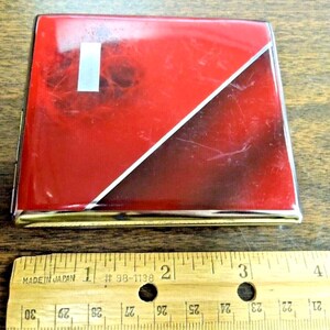 May include: A red and black cigarette case with a gold trim. The case has a silver stripe running diagonally across the front. The case is about 3 inches long.