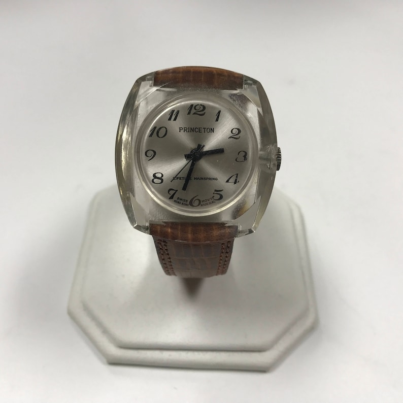 Peut inclure: Une montre-bracelet en cuir marron avec un bo&icirc;tier en plastique transparent. Le cadran de la montre est blanc avec des chiffres et des aiguilles noirs. Le cadran de la montre comporte &eacute;galement le texte "PRINCETON" et "SWISS MOVEMENT".