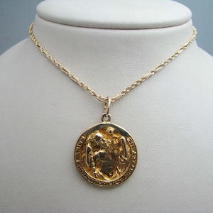 May include: A gold chain necklace with a round pendant featuring a detailed relief of a winged figure. The pendant has an inscription around the edge.