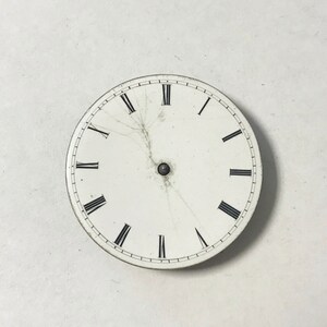 Puede incluir: Una esfera de reloj blanca con marcas de n&uacute;meros romanos negros y un peque&ntilde;o punto negro en el centro. La esfera del reloj tiene varias grietas que irradian desde el centro.