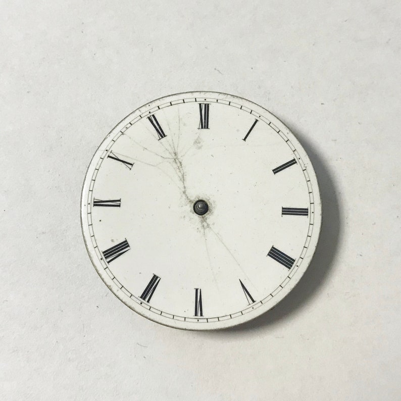 Puede incluir: Una esfera de reloj blanca con marcas de n&uacute;meros romanos negros y un peque&ntilde;o punto negro en el centro. La esfera del reloj tiene varias grietas que irradian desde el centro.