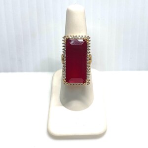 May include: A gold-toned ring featuring a large, rectangular, deep red gemstone. The gemstone is surrounded by a frame of small, clear stones and gold-colored accents. The ring is displayed on a white stand against a white background.