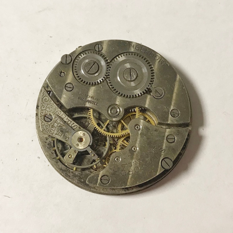 May include: Close-up of a silver pocket watch movement with the inscription "LANGENDORF" and "SUISSE" on the face. The watch has a variety of gears and springs visible.
