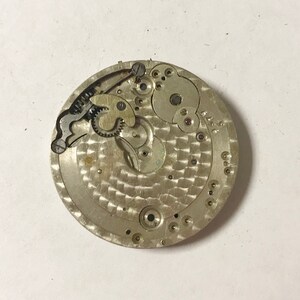 Puede incluir: Primer plano de un movimiento de reloj plateado con engranajes intrincados y una superficie texturizada. El movimiento carece de manecillas y tiene un peque&ntilde;o agujero en el centro.