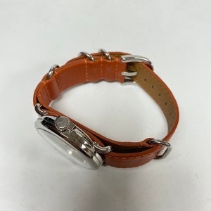 May include: A silver-toned wristwatch with a light brown leather band. The watch face is round with a silver bezel. The band features silver metal loops and a buckle closure. The watch is a classic accessory.