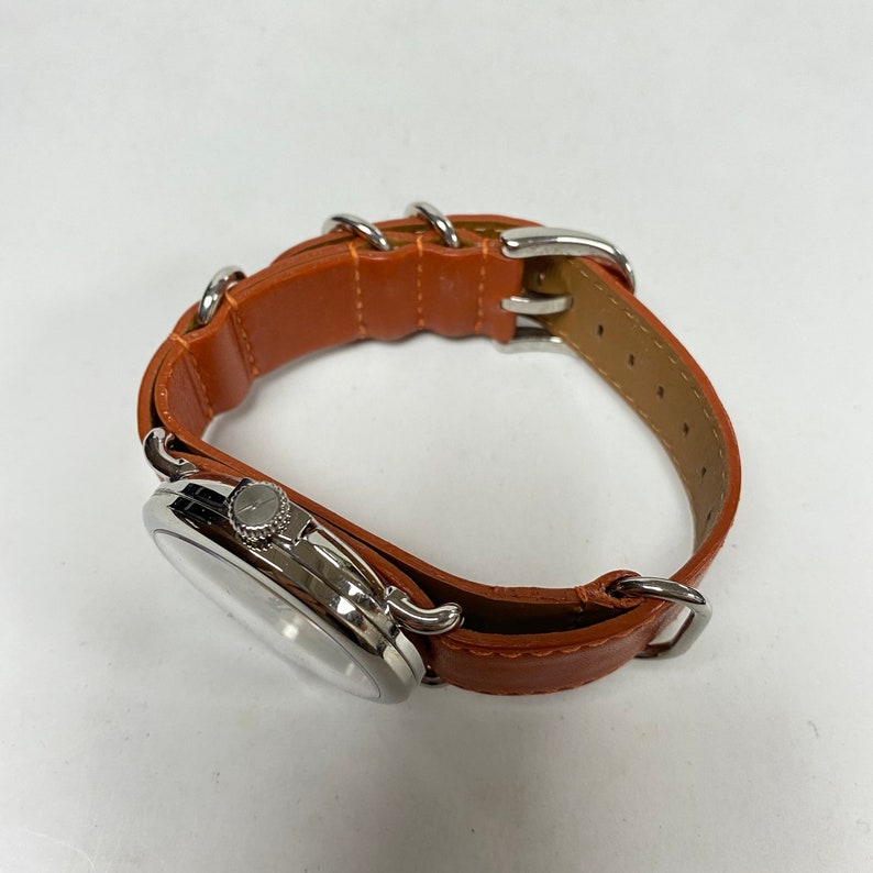 May include: A silver-toned wristwatch with a light brown leather band. The watch face is round with a silver bezel. The band features silver metal loops and a buckle closure. The watch is a classic accessory.