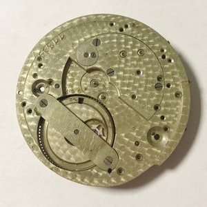 Pu&ograve; includere: Primo piano di un movimento di orologio argentato con ingranaggi intricati e una superficie testurizzata. Il movimento &egrave; etichettato "17914".