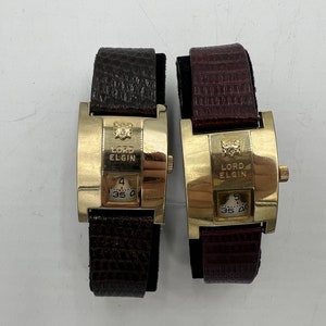 May include: Two gold-tone wristwatches with brown and burgundy leather bands. Both watches feature a square face with a white dial and the text "Lord Elgin".