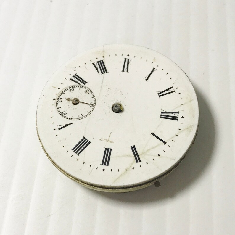 Puede incluir: Esfera de reloj de bolsillo blanca con marcadores de hora en n&uacute;meros romanos y un dial de segundero circular m&aacute;s peque&ntilde;o. La esfera del reloj tiene un aspecto desgastado y envejecido con ara&ntilde;azos visibles.
