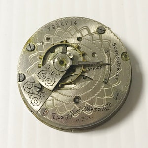 Op de afbeelding: Een zilveren zakhorloge met een witte wijzerplaat en zwarte markeringen. De wijzerplaat van het horloge heeft een decoratief ontwerp en de tekst "Elgin Natl Watch Co. U.S.A." en "Seven Jewels". De wijzerplaat van het horloge heeft ook het nummer "7926714".