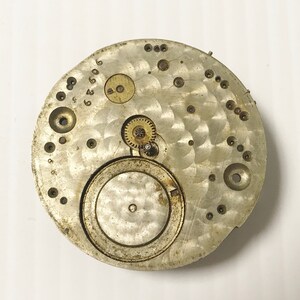 May include: Close-up of a silver watch movement with exposed gears and a circular dial. The movement is marked with numbers and has a worn, aged appearance.