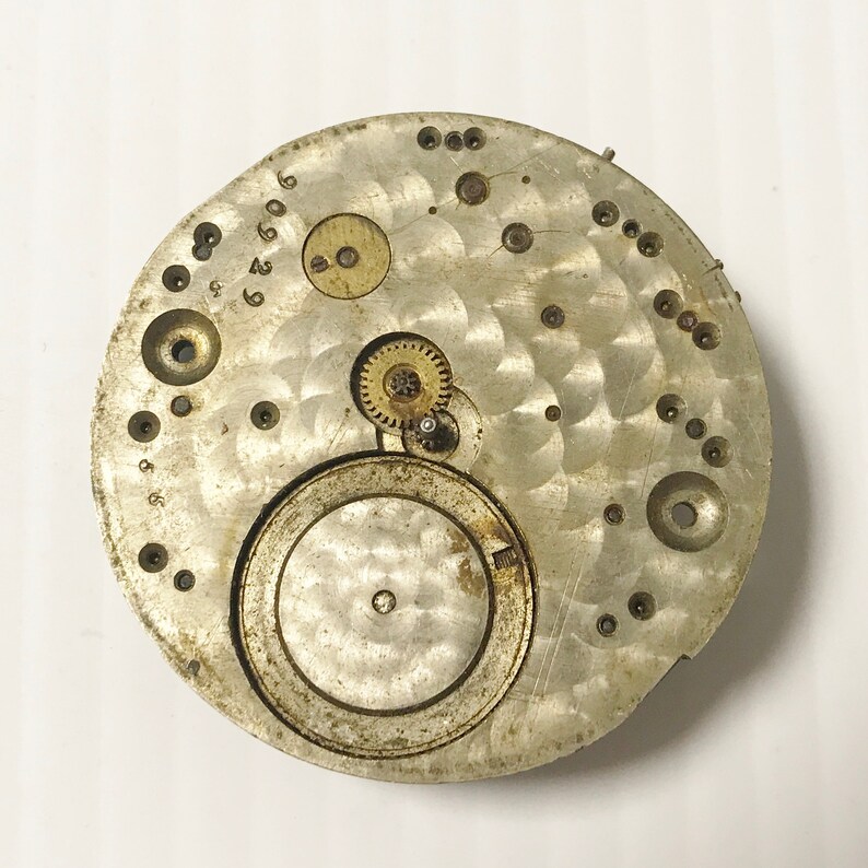 May include: Close-up of a silver watch movement with exposed gears and a circular dial. The movement is marked with numbers and has a worn, aged appearance.