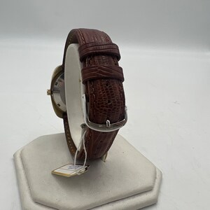 Puede incluir: Una correa de reloj de cuero marr&oacute;n con una hebilla plateada. La correa est&aacute; en un expositor blanco.