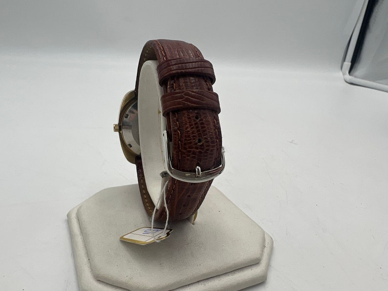 Puede incluir: Una correa de reloj de cuero marr&oacute;n con una hebilla plateada. La correa est&aacute; en un expositor blanco.