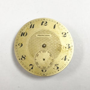 Puede incluir: Una esfera de reloj vintage dorada con n&uacute;meros negros y una subesfera. La palabra "PRINCIPAL" est&aacute; impresa encima del centro. La palabra "SWISS" est&aacute; impresa en la parte inferior. La esfera tiene un centro texturizado.